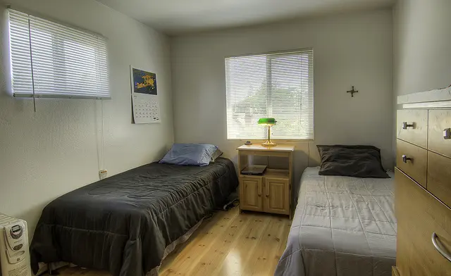 Interior bedroom showing two twin beds, nightstand, windows with blinds, and hardwood flooring