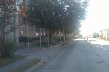 Side exterior of brick facility building with security fencing, mature trees, and paved street frontage.