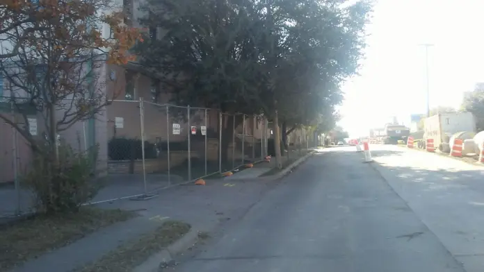 Side exterior of brick facility building with security fencing, mature trees, and paved street frontage.