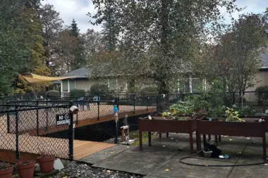 Outdoor recreational space with raised garden beds, mature trees, seating tables, and landscaping