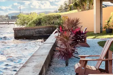 Outdoor patio seating area with Adirondack chairs, palm trees, landscaping, concrete seawall, and waterfront river views.