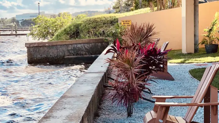 Outdoor patio seating area with Adirondack chairs, palm trees, landscaping, concrete seawall, and waterfront river views.