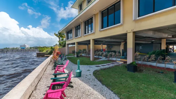 Exterior patio seating with pink Adirondack chairs, landscaping, grass, pathways, and river view near yellow building.